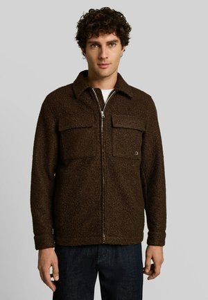 Junger Mann mit lockigem Haar, der eine dunkelbraune, strukturierte Reißverschlussjacke mit aufgesetzten Brusttaschen und dunkle Hosen vor einfachem Hintergrund trägt.