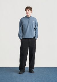 Ανοιχτό μπλε, φούτερ με φερμουάρ στον γιακά από μαλακό ύφασμα, συνδυασμένο με φαρδιά μαύρη παντελόνια cargo με πολλές τσέπες. Το μοντέλο φοράει μαύρα αθλητικά παπούτσια.