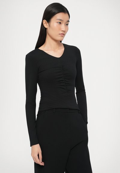 Νεαρή γυναίκα με μακριά μαύρα μαλλιά, ντυμένη με εφαρμοστό μαύρο τοπ με μακριά μανίκια και ρυτιδωμένη μπροστινή όψη, καθώς και μαύρο παντελόνι, ποζάρει μπροστά σε απλό φόντο.