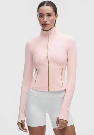 Mujer con una chaqueta rosa claro con cremallera y agujeros para los pulgares, y pantalones cortos deportivos blancos de talle alto, sobre un fondo liso.
