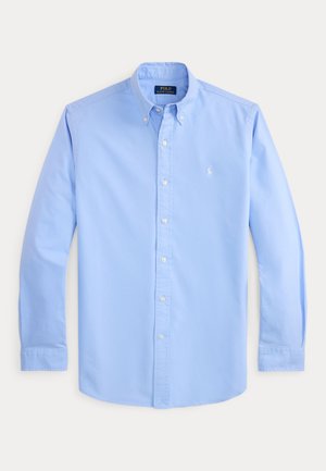 Chemise bleu clair à manches longues avec col boutonné, boutons blancs et petit logo blanc brodé sur la poitrine gauche.