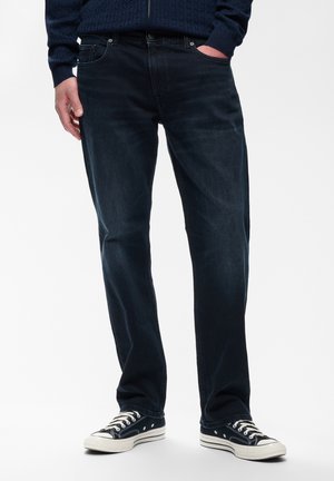 Homme portant un jean bleu foncé, un pull bleu marine et des baskets basses noires et blanches, debout avec une main dans la poche.