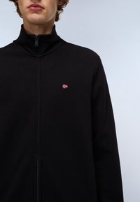 Schwarze Zip-Jacke, glattes Material, hoher Kragen, mit einer kleinen, gestickten norwegischen Flagge auf der linken Brust. Einfaches Design, keine Muster.