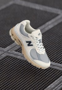 Beige and gray New Balance sneaker with white laces and tan sole, placed on an industrial metal grate surface.