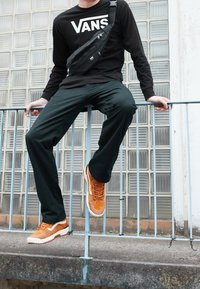 Person in schwarzem Vans-Shirt und -Hose mit orangen Vans-Sneakern und Umhängetasche, der auf einem Metallgeländer in städtischer Umgebung sitzt.