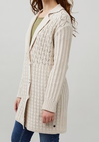 Cardigan en tricot beige clair avec un motif en losanges texturé, doté d'un col châle, d'une fermeture à boutons et de poignets côtelés.