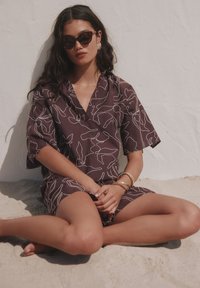 Femme portant un haut court à manches courtes foncé à motifs et un short, des lunettes de soleil et des bijoux en or, assise en tailleur sur un sol sablonneux contre un mur blanc.