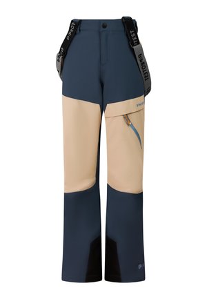 Een jong persoon staat en draagt een beige fleecejack, een oranje muts, blauwe en beige sneeuwbroek met bretels en zwarte winterlaarzen op een effen achtergrond.