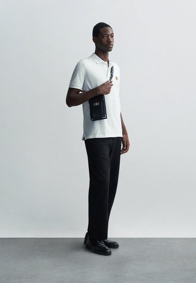Tricou polo alb cu un mic detaliu brodat, pantaloni negri și pantofi negri. Persoana ține o mică geantă de umăr neagră.