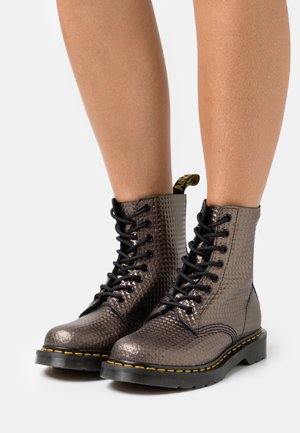 Personne portant des bottines à lacets avec une texture métallique, semelles noires et surpiqûres jaunes sur un fond blanc.