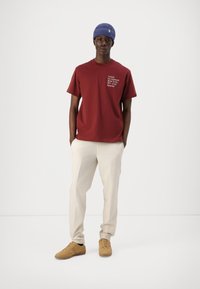 Burgunder bomull T-skjorte med hvit tekst på forsiden, parret med lyse beige bukser og brune sneakers. Modellen har på seg en blå strikket lue.