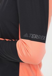 Camicia a maniche lunghe nera e corallo realizzata in materiale traspirante, con un motivo testurizzato. Presenta il logo "adidas TERREX" sulla manica.