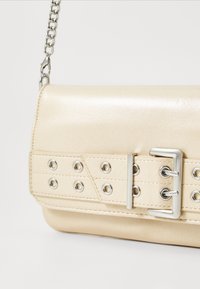 Sac à main en cuir beige avec une large ceinture et une boucle en argent, comportant des œillets argentés et une bandoulière en chaîne argentée.