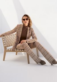 Woven plaid suit with neutral tones; tapered trousers, fitted blazer, ribbed brown turtleneck, and chunky heeled shoes. Wooden chair.