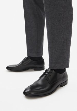 Czarne skórzane buty wizytowe z zaostrzonym noskiem, posiadające cztery pary dziurek na sznurówki, gładką fakturę, noszone z szarymi spodniami w wzory.