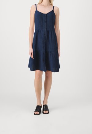 Femme portant une robe bleu marine sans manches à boutons sur le devant avec des volants plissés et des mules noires à bout ouvert, sur un fond clair uni.