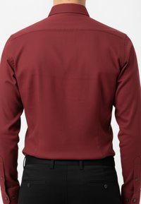 Hombre con una camisa de dress de manga larga ajustada de color rojo oscuro metida en pantalones de vestir negros, visto desde atrás contra un fondo blanco.