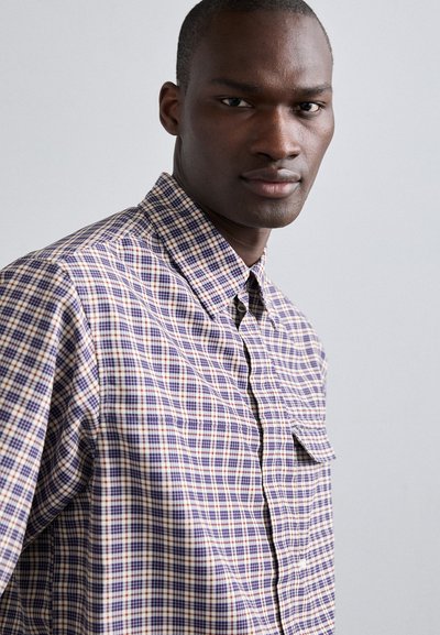 Camisa de cuadros que presenta una combinación de colores púrpura, rojo y crema. Fabricada en tejido de punto con cuello de botón y detalle de bolsillo en la parte frontal.