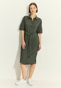 Vestito camicia verde oliva, lunghezza al ginocchio, con maniche corte, colletto, apertura frontale con bottoni e cintura in vita. Indossato con sneakers bianche, visibili fessure laterali.