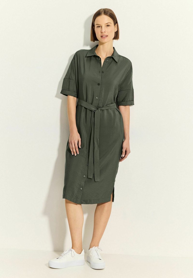 Vestito camicia verde oliva, lunghezza al ginocchio, con maniche corte, colletto, apertura frontale con bottoni e cintura in vita. Indossato con sneakers bianche, visibili fessure laterali.