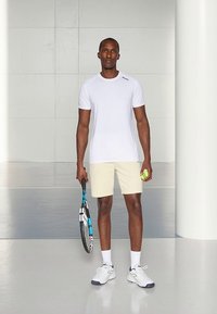 Camiseta desportiva branca, calções creme, segurando uma raquete de ténis e uma bola de ténis amarelo néon. Tecido macio, corte relaxado, detalhes contrastantes.