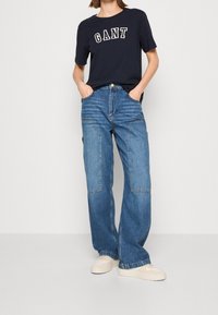 Mariene T-shirt met "GANT" tekst; losse lichtblauwe jeans met patchworkontwerp; gecombineerd met beige sneakers; zachte stof, casual stijl.