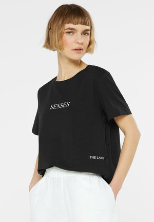 Young woman with short blond hair wears a black T-shirt with "SENSES" and "THE LABEL" text, paired with white pants, hands in pockets.