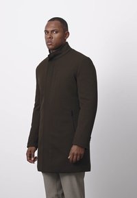 Hnědý vlněný kabát s vysokým límcem, předním zipem, dvěma bočními kapsami na zip a vypasovaným střihem. Hladká textura, krejčovský design.
