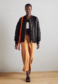Schwarze Bomberjacke, weißes Crop-Top, fließender orangener Rock, schwarze Rutschsandalen, eine orange Umhängetasche tragend. Strukturierte Stoffe, kontrastierende Farben.