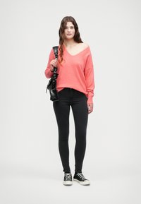 Suéter de punto coral de gran tamaño con cuello en V, llevado sobre unos pantalones vaqueros skinny negros y zapatillas de lona negras; sostiene un pequeño bolso cruzado negro.