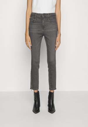 Osoba má na sobě šedé úzké džíny s detaily zipů u kotníků, doplněné černými kotníkovými botami s ostrou špičkou a bílým tílkem bez rukávů.