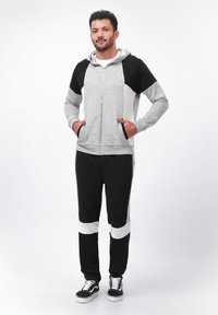 Grijze rits hoodie met zwart-witte accenten, gecombineerd met zwarte joggingbroek met horizontale witte strepen. Model draagt sneakers.
