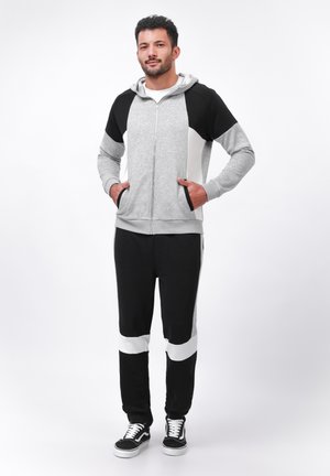 Grijze rits hoodie met zwart-witte accenten, gecombineerd met zwarte joggingbroek met horizontale witte strepen. Model draagt sneakers.