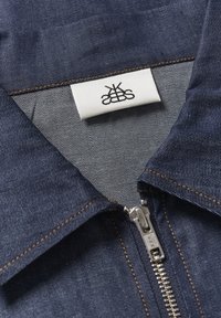 Dżinsowa kurtka w ciemnoniebieskim kolorze, z metalowym suwakiem, beżowymi szwami i białą metką z czarnym logo wewnątrz kołnierza.