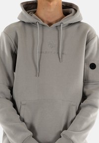 Felpa grigia realizzata in tessuto morbido, con tasca a marsupio, cappuccio regolabile con cordino e branding ricamato sul petto e sulla manica.