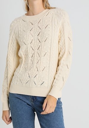 Femme portant un pull en maille torsadée crème avec des motifs de feuilles et de boules, associé à un jean bleu, debout devant un fond uni.