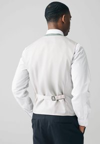Light-colored vest with a smooth texture, featuring a back strap and buckle detail, paired with a white shirt and dark trousers.