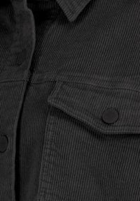 Schwarzer Cordstoff mit sichtbarer gerippter Textur, mit einem Kragen und zwei Vordertaschen mit runden schwarzen Knöpfen.