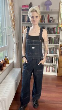 Dark denim overalls with wide legs and adjustable straps. Front pocket and side pockets visible. Black top underneath, minimalistic design.