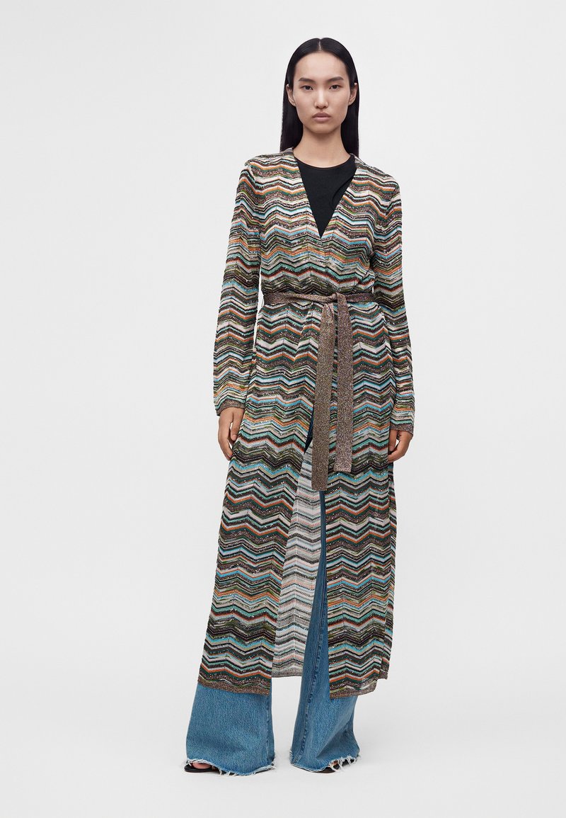 Femme portant une longue robe portefeuille multicolore à motif zigzag sur un top noir et un jean bleu à jambes larges, debout devant un fond uni.