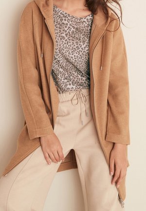 Femme portant un long sweat à capuche camel zippé, un haut léopard et un pantalon beige à cordon, debout avec une main posée sur la cuisse.