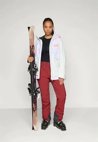 Skikleding bestaande uit een lichtblauwe, hoodie-jack met grafische accenten, bordeauxrode skibroeken en zwarte skiskimmen. Ski's worden aan de zijkant vastgehouden.