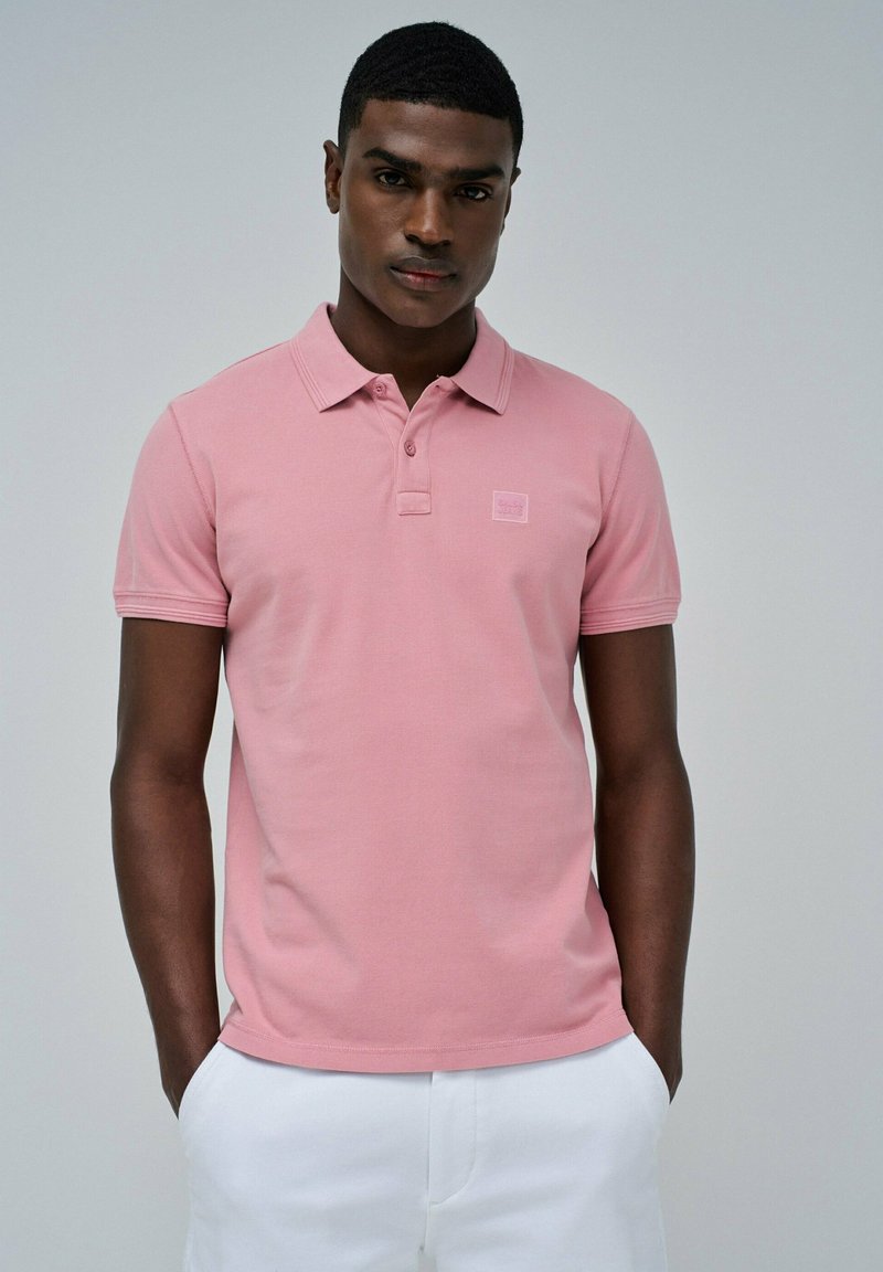Polo de color rosa claro hecho de tela texturizada, con cuello y tapillera de dos botones, combinado con pantalones cortos blancos. Sin patrones notables.