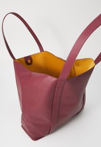 Maroon leather tote bag with yellow lining, featuring two long handles and a snap button closure. Smooth texture with minimalist design.