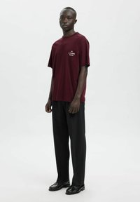 Burgundska bombažna majica s kratkimi rokavi in okrolim izrezom, z belim napisom "LA FLAMME ROUGE" na levi strani prsnega koša. Nošena s črnimi hlačami.