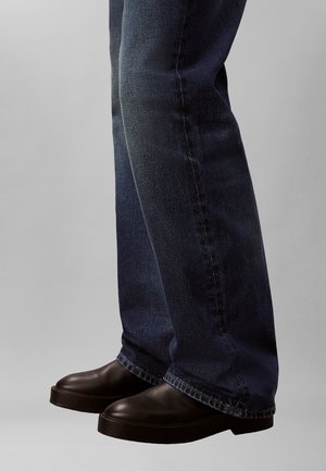 Scarpe nere slip-on con punta arrotondata e suola spessa, abbinate a jeans denim blu scuro con vestibilità rilassata e leggera scoloritura.