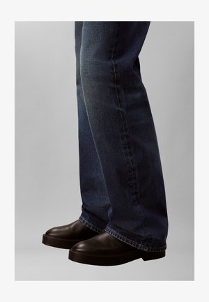 Zapatos negros sin cordones con punta redonda y suela gruesa, combinados con vaqueros de mezclilla azul oscuro que muestran un ajuste relajado y un ligero desgaste.