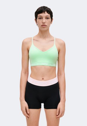 Mujer con cabello corto y oscuro que lleva un sujetador deportivo Nike verde claro y pantalones cortos negros de talle alto con una cintura de color rosa pálido.