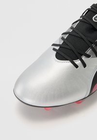 Botte de football synthétique argentée avec un bout arrondi, une zone de lacet en mesh noir et des crampons avec des accents roses sur la semelle. Texture lisse, design aérodynamique.