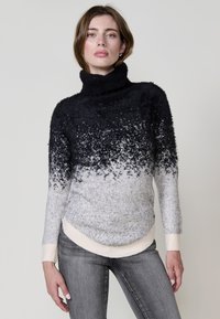 Črno-sivo ombre turtleneck pulover iz mehkega, puhastnega materiala z rebrastimi manšetami in ukrivljenim robom. Obut je z sivimi kavbojkami.
